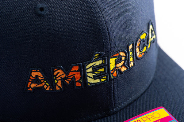 Club America Day of the Dead Snapback Hat with America script with marigold floral overlay embroidered on the front, Club America logo embroidered on the side, "Dia de Muertos" embroidered on the back, and marigold floral print on the under peak. The hat has a high crown, flat peak brim, and a snapback closure.