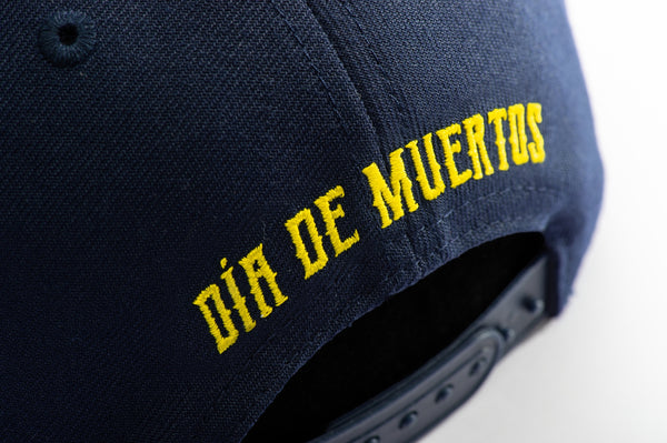 Club America Day of the Dead Snapback Hat with America script with marigold floral overlay embroidered on the front, Club America logo embroidered on the side, "Dia de Muertos" embroidered on the back, and marigold floral print on the under peak. The hat has a high crown, flat peak brim, and a snapback closure.